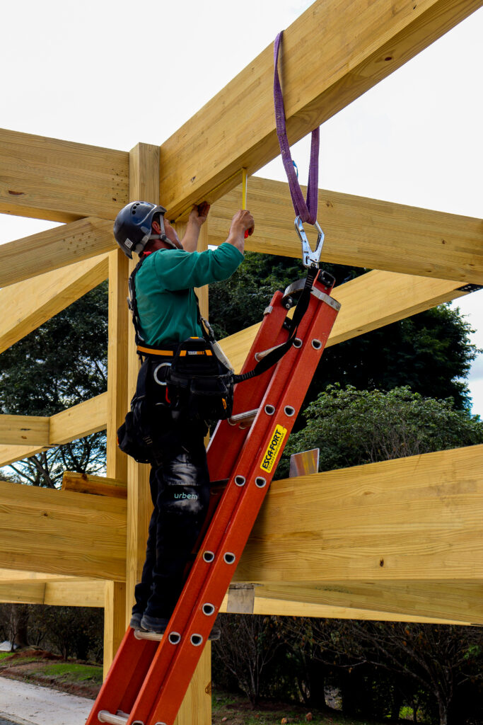 Montador da Urbem em pé sobre uma escada de fibra, utilizando equipamentos de proteção individual e ferramentas presas ao cinto, enquanto mede e fixa peças de uma estrutura de madeira engenheirada em montagem a céu aberto.